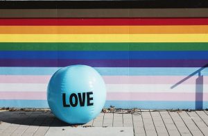 A beach ball with the word Love sits in the foreground of a wall painted with 2SLGBTQIA rainbow stripes.